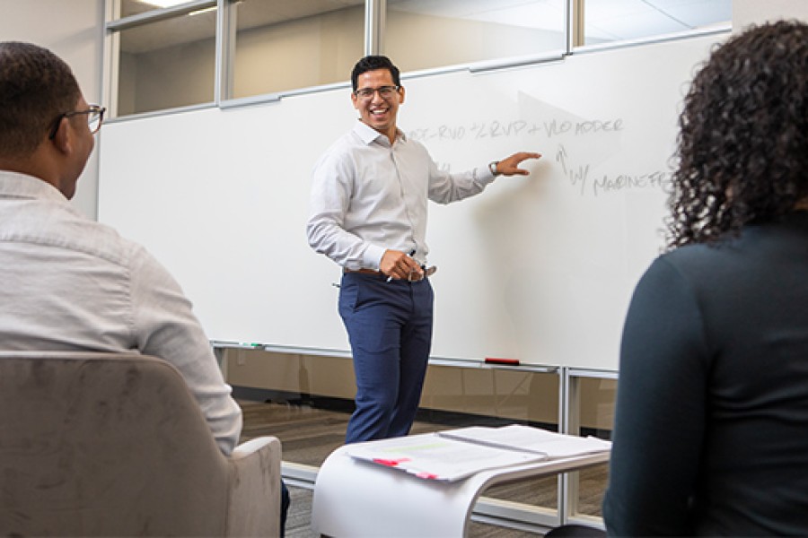 a man and a woman seated, another man smiling and pointing to whiteboard 